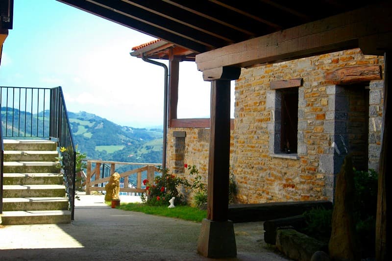 Porche de piedra y madera del caserío, con rosales y vistas abiertas al valle