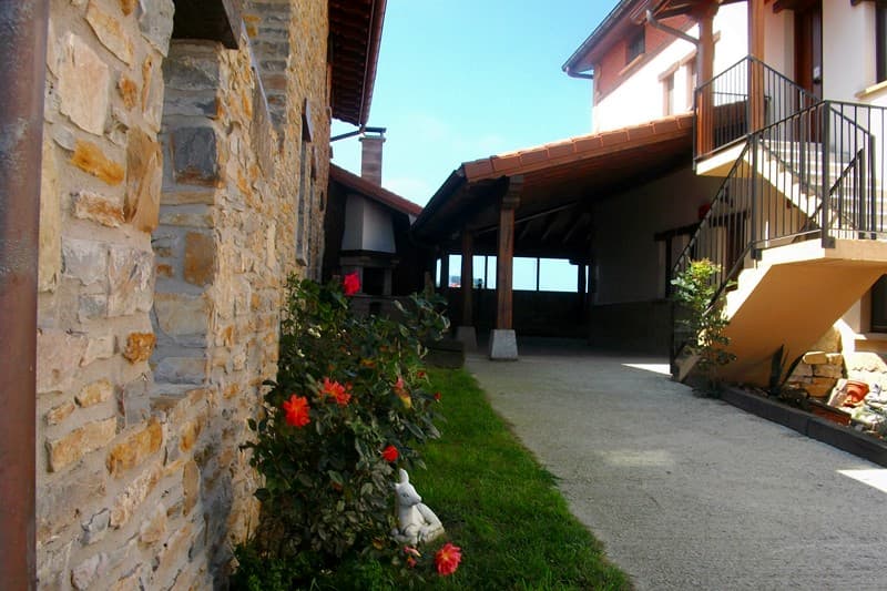 Camino de grava junto al muro de piedra del caserío, con rosales y escalera exterior