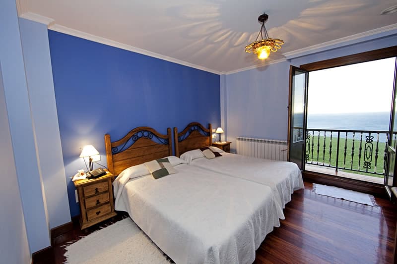 Habitación de paredes azules con dos camas, colchas blancas y balcón abierto al mar Cantábrico